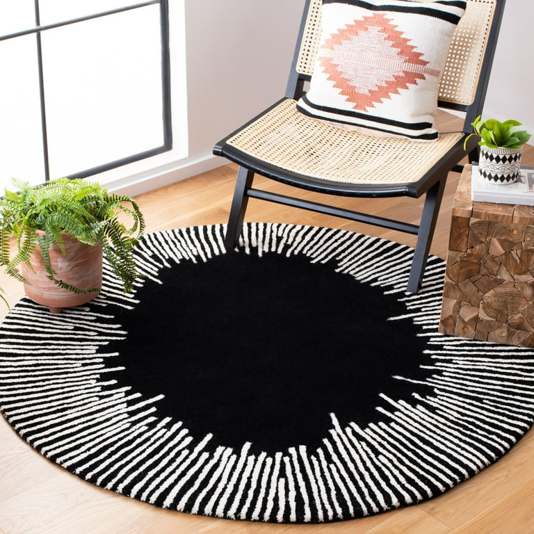 Modern round black area rug with a white sunburst stripe pattern, styled in a living room under a black woven chair and next to a wooden side table.