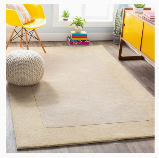 Rectangular solid beige rug with a tonal cream border in a modern, sunlit living room.